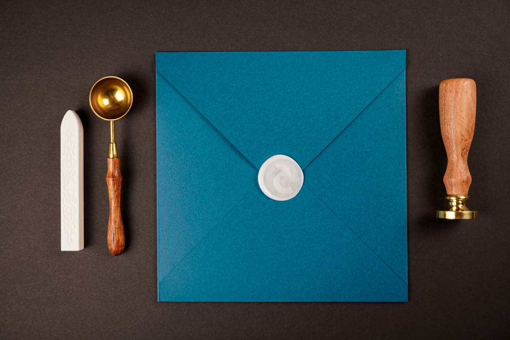 Flatlay Of A Petrol-colored Envelope With Wax Seal, To The Left There Is A White Sealing Wax Stick And The Spoon For Melting The Wax, To The Right There Is The Sealing Stamp, Part Of The Brand Identity Design For Corliss Design