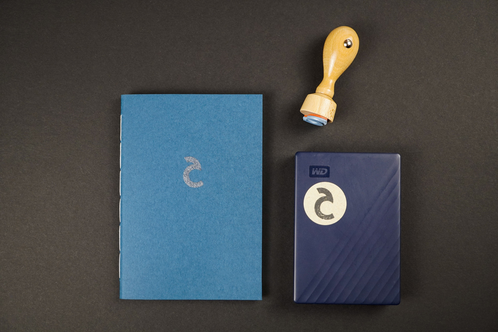 Flatlay Of A Hand-bound Booklet With Logo Embossing On Cover And External Hard Drive With Logo Sticker On With The Stamp As Part Of The Brand Identity Design For Corliss Design